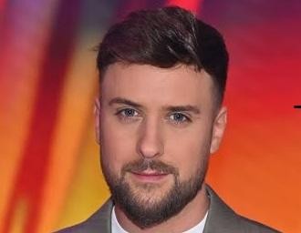 Aleksander Tomasz Sikora, prezenter telewizyjny, w swobodnej pozycji, przeglądający media społecznościowe na telefonie, uśmiechający się do obiektywu, symbolizujący jego interakcję z fanami online.