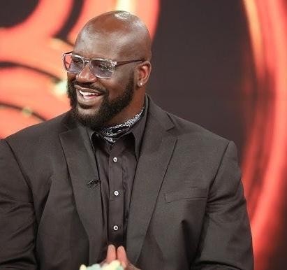 Shaquille O'Neal w garniturze trzymający dyplom na tle półki z książkami, symbolizujący jego osiągnięcia edukacyjne.