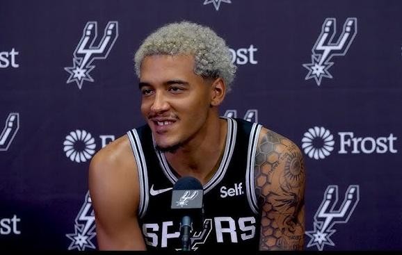 Jeremy Sochan w zbliżeniu, w stroju San Antonio Spurs, z charakterystycznymi włosami, skupiony podczas treningu lub rozgrzewki.