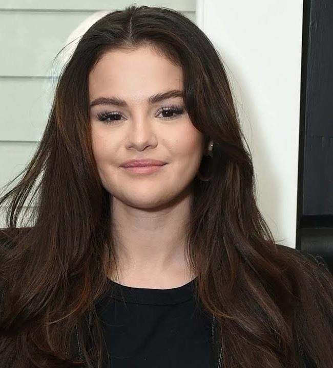 Selena Gomez na scenie podczas koncertu, śpiewająca do mikrofonu, z oświetleniem scenicznym i tłumem w tle.