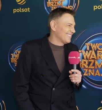 Krzysztof Ibisz w studiu telewizyjnym, dynamicznie prezentujący program, ubrany w elegancki garnitur, z charyzmatycznym wyrazem twarzy.